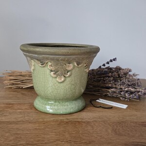 May include: A light green ceramic planter with a decorative rim and base. The planter has a crackle finish and is displayed on a wooden surface. Dried lavender and a bundle of straw are next to the planter.