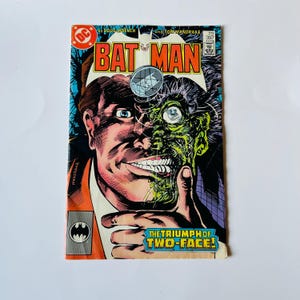 May include: A vintage Batman comic book featuring a split-faced character, Two-Face, with one side normal and the other disfigured. The cover has the title "BATMAN" in bold orange letters and the text "THE TRIUMPH OF TWO-FACE!"