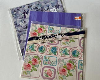 May include: Three packages of folded gift wrap. The top sheet features a floral pattern with pink hibiscus and blue morning glories in rectangular frames, labeled All-Occasion Gift Wrap. Beneath it is a purple striped sheet and a blue hydrangea print.