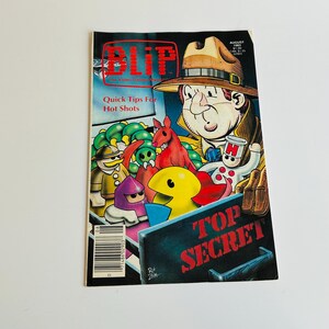 May include: A magazine cover featuring a cartoon illustration of a man in a fedora and trench coat, holding a sign that says "Top Secret". The magazine is called "Blip" and the cover text says "Quick Tips For Hot Shots".