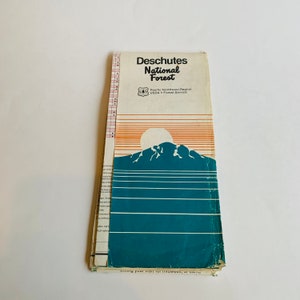 May include: A vintage map of the Deschutes National Forest in Oregon, featuring a graphic of a mountain range with a sun rising behind it. The map is printed on white paper with orange and blue accents. The text "Deschutes National Forest" is printed in black at the top of the map. The text "Pacific Northwest Region USDA Forest Service" is printed in black below the title.