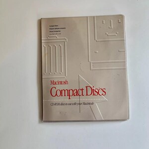 May include: A beige Macintosh Compact Discs case. The case has embossed architectural designs and text in English, French, Spanish, and Japanese. The red text reads "Compact Discs" and "CD-ROM discs to use with your Macintosh."