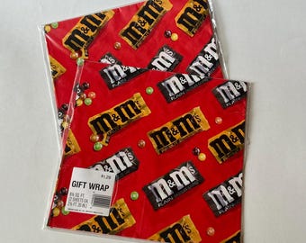 May include: Two folded sheets of red gift wrap featuring a repeating pattern of M&M's chocolate candy packages and individual colorful candies. A white product label is attached to the front, stating GIFT WRAP, 8 1/2 SQ. FT., and 2 1/2 FT. 20 IN.