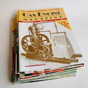 May include: A stack of vintage Gas Engine Magazine issues. The top magazine has a yellow and white cover with the title "Gas Engine Magazine" and an illustration of an antique engine. The magazine's website is visible.