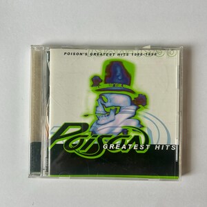 May include: A CD case featuring the album "Poison's Greatest Hits 1986-1996." The cover art displays a skull wearing a hat, with the band's logo in green. The CD case is clear, and the background is a light green and black.