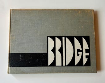 May include: A rectangular hardcover book with a light gray fabric cover. A horizontal black stripe runs across the lower portion, featuring the word BRIDGE in large, bold, white capital letters with a stylized, geometric, and curved font design.