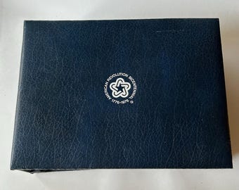 May include: A rectangular dark blue box with a textured, leather-like finish. Centered on the lid is a white circular logo featuring a star design and the text "AMERICAN REVOLUTION BICENTENNIAL 1776-1976" printed in a clean, sans-serif font.