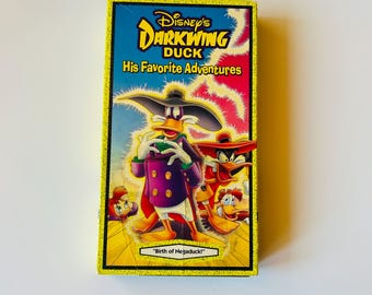 May include: A rectangular VHS tape case featuring Disney's Darkwing Duck His Favorite Adventures. The cover art displays the purple-clad character Darkwing Duck in a wide-brimmed hat, with other duck characters, against a yellow and pink background.