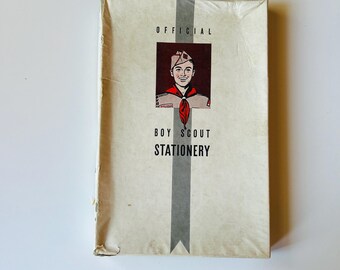 May include: A rectangular off-white stationery box featuring a central gray vertical stripe and a graphic illustration of a scout wearing a tan uniform and a red neckerchief. The text reads OFFICIAL BOY SCOUT STATIONERY in black lettering.