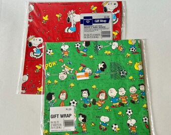 May include: Two packages of Ambassador gift wrap. The red sheet features Snoopy and Woodstock with musical notes. The green sheet displays Peanuts characters playing soccer with the text POW. Each package includes two sheets measuring 2.5 ft by 20 in.