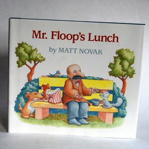 May include: A children's book cover with a cartoon illustration of a man named Mr. Floop sitting on a park bench with a squirrel and a dog. The book title is "Mr. Floop's Lunch" by Matt Novak.
