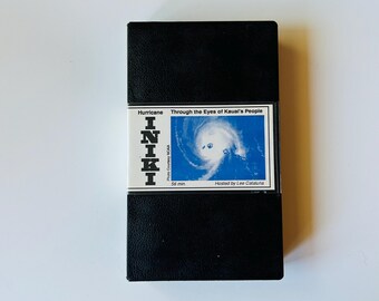 May include: A black VHS cassette tape with a white label featuring the text Hurricane Iniki Through the Eyes of Kauai's People, a blue satellite image of a hurricane, the text 56 min, Photo Courtesy NOAA, and Hosted by Lee Catalina.
