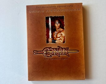 May include: A brown rectangular DVD box set titled The Franchise Collection Arnold Schwarzenegger Conan The Complete Quest. The cover features a central image of a muscular figure holding a sword, with the title text displayed below.