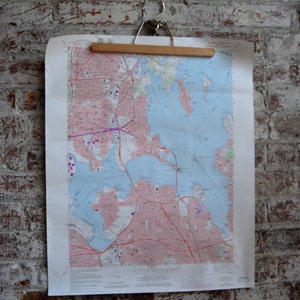 Puede incluir: Un mapa de Boston, Massachusetts, colgado en una pared de ladrillo. El mapa está impreso en papel blanco y muestra las calles, vías fluviales y puntos de referencia de la ciudad. El mapa está sujeto a un colgador de madera.