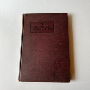 May include: A vintage book with a dark burgundy cover. The title "The Automatic Letter Writer" is printed in a rectangular frame on the front. A small emblem is visible near the bottom right corner.