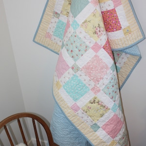 Könnte beinhalten: Eine pastellblaue, rosafarbene, gelbe und weiße Patchwork-Decke mit Blumenmuster hängt an einem Holzhaken. Ein weißer Teddybär sitzt auf einem Holz-Schaukelstuhl.