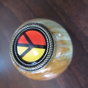 May include: A wooden knob with a metal ring around a colorful peace sign. The peace sign is red, yellow, and black with a black outline.