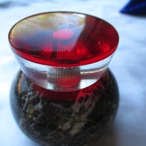 May include: A red glass bottle stopper with a clear base sits on a black and white marble base.