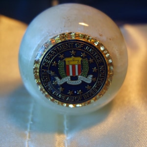 May include: A round, off-white decorative object with a gold-rimmed emblem featuring the text "DEPARTMENT OF JUSTICE FEDERAL BUREAU OF INVESTIGATION" and a central design. The emblem is blue, gold, and red.