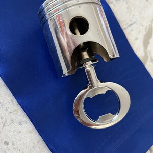 May include: A silver-colored metal bottle opener featuring a cylindrical engine piston head design. The top portion is a smooth, polished piston with horizontal grooves, while the bottom portion functions as a standard loop-style bottle opener.