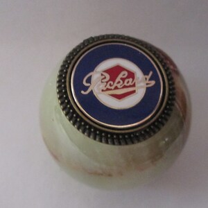 May include: A decorative, round object with a green and brown marbled appearance. The top features a blue circle with the word "Packard" in white script, surrounded by a gold-colored metal trim.