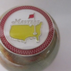 May include: A golf ball marker with a white face featuring the word "Masters" and a yellow outline of the United States with a golf flag. The marker has a red and white border and a multi-colored base.