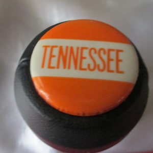 May include: A black bottle stopper with an orange and white round top that says "TENNESSEE" in white letters.