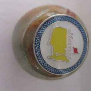 May include: A green and brown marble paperweight with a silver and blue circular design featuring the Masters Tournament logo. The logo is yellow and white with a red flag.