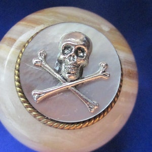 May include: A round, white, and brown marble orb with a silver skull and crossbones design on a silver and gold circular inlay.