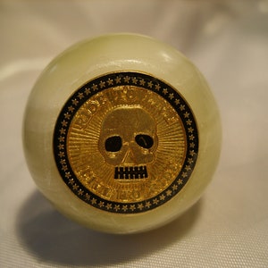 May include: A white marble sphere with a gold skull and crossbones design. The skull is surrounded by a black circle with gold stars.