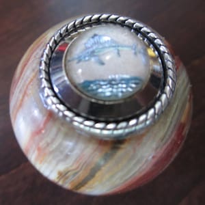May include: A round, multi-colored, marble-like knob with a silver-colored, rope-like trim. The knob has a clear, round, glass insert with a painted image of a marlin fish jumping out of the water.