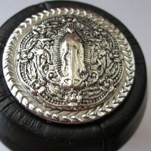 May include: A silver-toned, round, decorative button with a raised image of a religious figure surrounded by intricate floral designs. The button is mounted on a black, leather-like base.