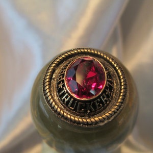 May include: A gold-colored ring with a red gemstone and the inscription "TRUCKER" around the stone.
