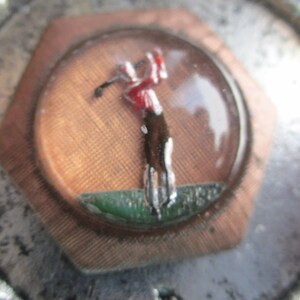May include: A silver and copper golf themed pin with a golfer in a red shirt and black pants swinging a golf club. The golfer is in a green field and the pin is surrounded by a silver rope design.