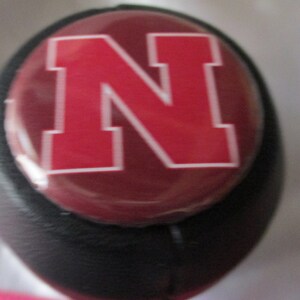 May include: A black gear shift knob with a red and white button featuring a large white outlined "N" in the center.