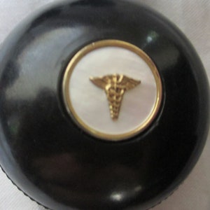 May include: A black round pill box with a gold rim and a white center featuring a gold medical symbol.