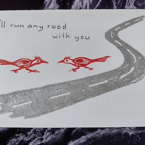 May include: A white card with red ink illustrations of two birds facing each other. The text "I'll run any road with you" is written in black ink.