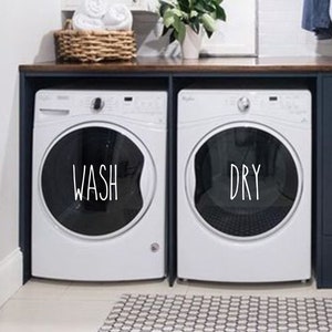 May include: A white washer and dryer with black labels that say "WASH" and "DRY" are in a laundry room with dark blue cabinets and a wood countertop.