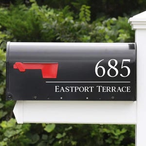 May include: A black mailbox mounted on a white post, featuring a red signal flag and white lettering that displays the number 685 above the text EASTPORT TERRACE. The mailbox has a curved top and is attached to a white horizontal support arm.