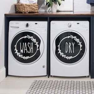 May include: White washing machine and dryer with black wreath decals that say "WASH" and "DRY" in a laundry room with dark blue cabinets and a white countertop.