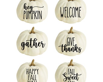 May include: Six white pumpkins with black lettering. The pumpkins are decorated with the following phrases: "hey pumpkin", "welcome", "gather", "give thanks", "happy fall y'all", and "fall sweet fall".