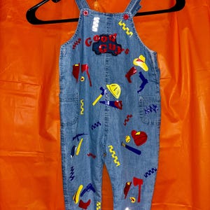 May include: Light blue denim overalls featuring red buttons and a variety of colorful patches including the text Good Guys, axes, hats, hammers, saws, baseball bats, and balls. The overalls are displayed on a black plastic hanger.