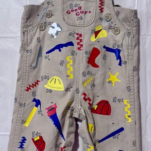 May include: Beige overalls featuring a repeating small black line-art pattern and larger colorful graphic prints of tools, hats, and weapons. The chest pocket displays the text Good Guys in red. The garment includes red buttons and metal hardware.