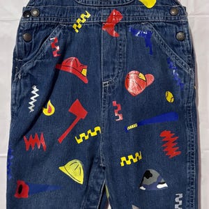 May include: Dark blue denim overalls featuring a red, yellow, and blue graphic print of various tools, hats, and shapes. The chest pocket displays a blue patch with the text Good Guys. The garment has red buttons and a plaid lining at the top.