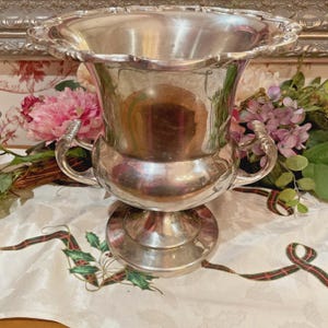 May include: A polished silver-toned metal wine cooler or ice bucket featuring a wide, scalloped rim, a rounded body, and two ornate, curved handles on opposite sides. The vessel stands on a circular pedestal base with a smooth, reflective finish.