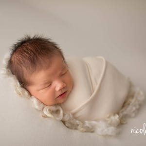 May include: A newborn baby is sleeping soundly, wrapped in a white swaddle blanket. The baby has dark hair and is lying on a white background.