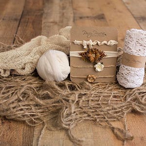 May include: Three brown cardboard cards with the text "Pretty Pieces" on the top card. Each card features a different floral headband with white and brown accents. The cards are displayed on a rustic wooden surface with a white crocheted headband, a white knitted ball, and a brown burlap fabric.