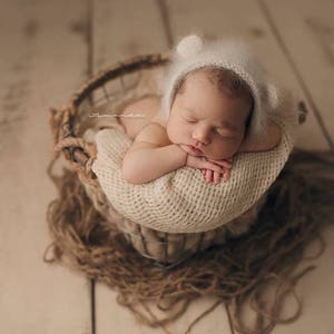 Puede incluir: Un bebé recién nacido con un gorro blanco peludo con orejas de oso está durmiendo en una cesta tejida forrada con una manta de color crema. La cesta está sobre una superficie de madera marrón claro.