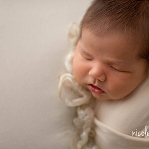 May include: A newborn baby is sleeping soundly, wrapped in a white swaddle with a delicate lace trim. The baby's face is peaceful and serene, with closed eyes and a soft expression.