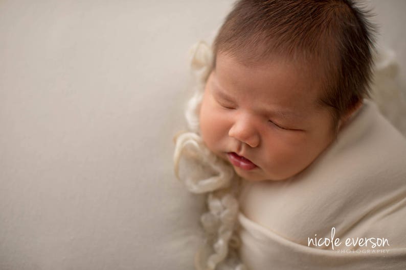 May include: A newborn baby is sleeping soundly, wrapped in a white swaddle with a delicate lace trim. The baby's face is peaceful and serene, with closed eyes and a soft expression.